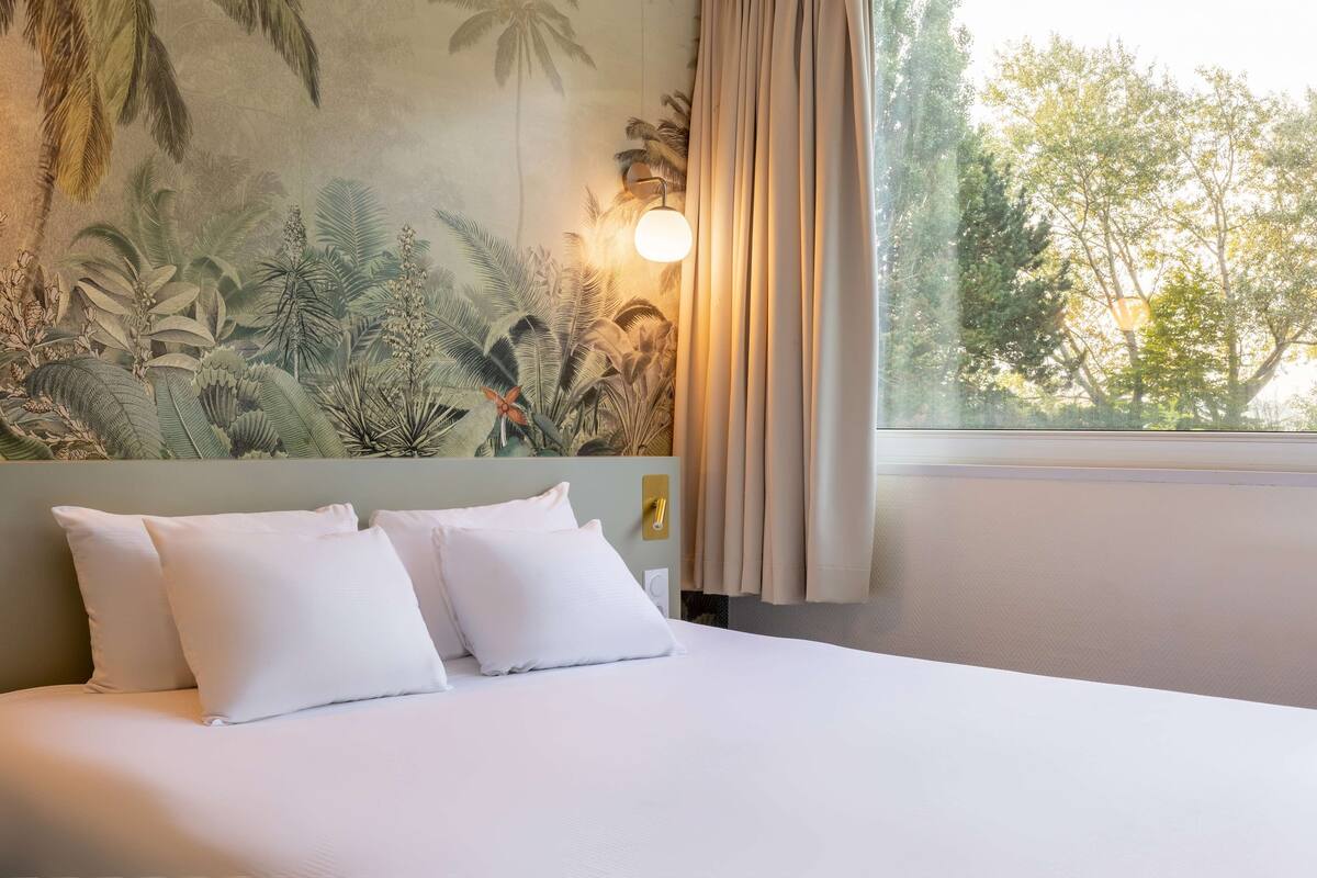 Une chambre d’hôtel avec un lit, des oreillers blancs, une fresque murale, une fenêtre avec des rideaux et un luminaire fixé au mur.