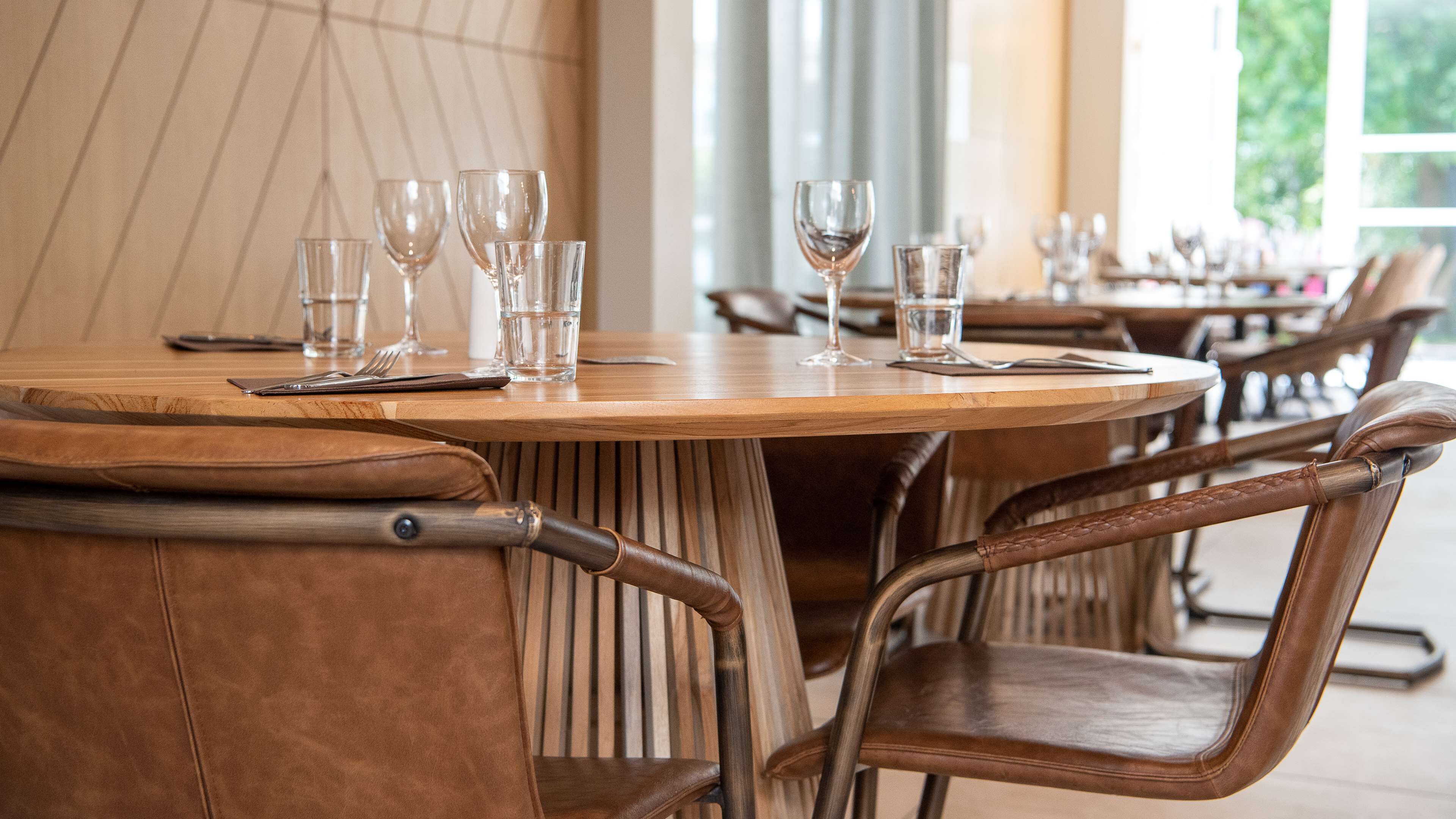 A restaurant interior with wooden tables and leather chairs, clear glassware, and a view of greenery outside.