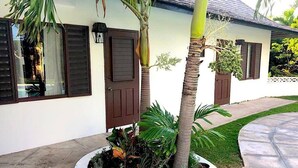 A house with a white exterior, dark shutters, and a circular garden bed with various plants and a palm tree.