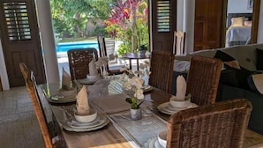 A dining area with a wooden table and wicker chairs, a vase with flowers, and a view of a swimming pool and greenery.