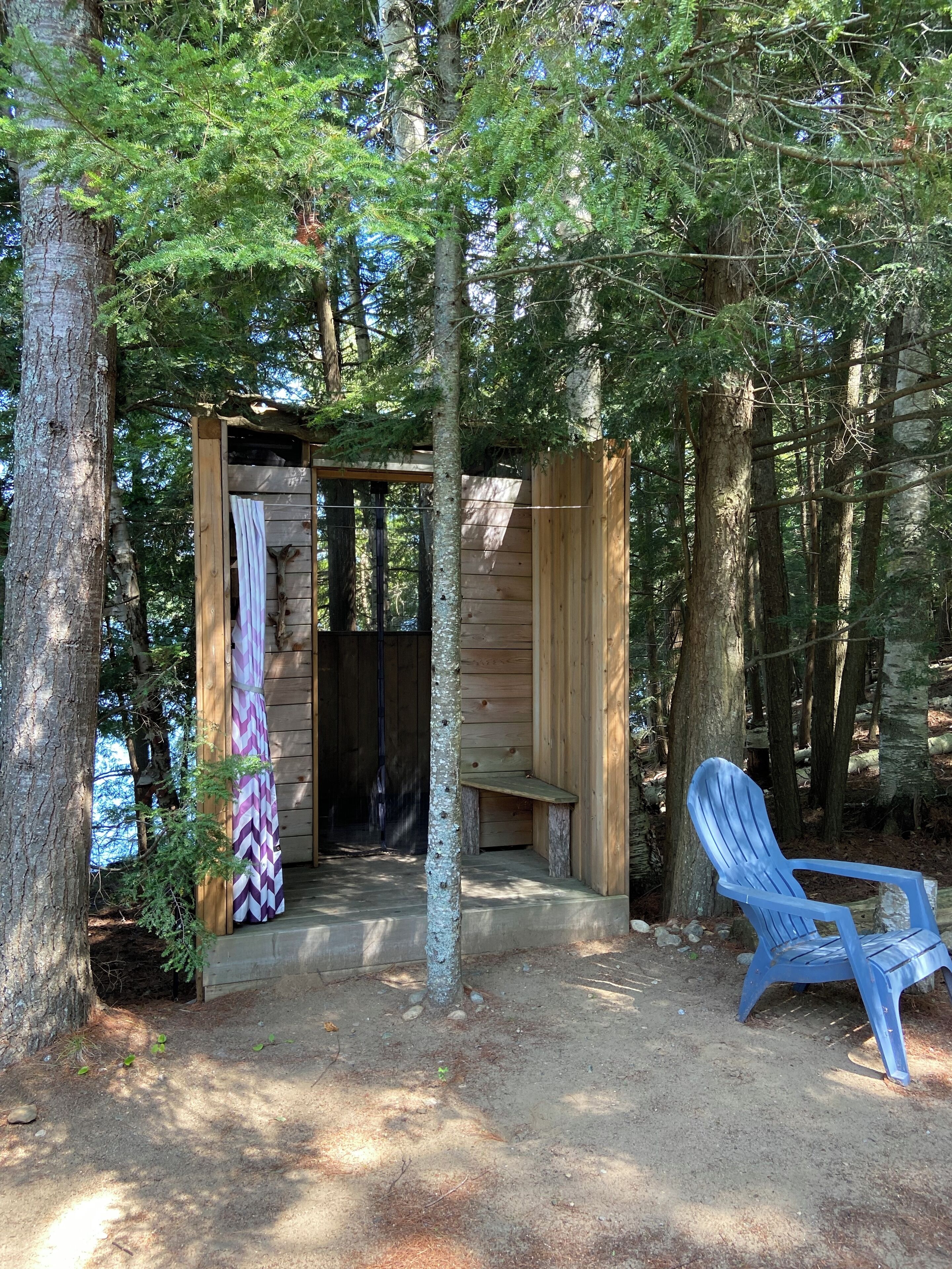 A wooden cabin with a bench and a chair outside, surrounded by trees.