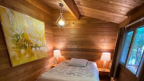 Een houten slaapkamer met een bed, twee bedlampjes, een schilderij aan de muur en een raam met uitzicht op bomen.