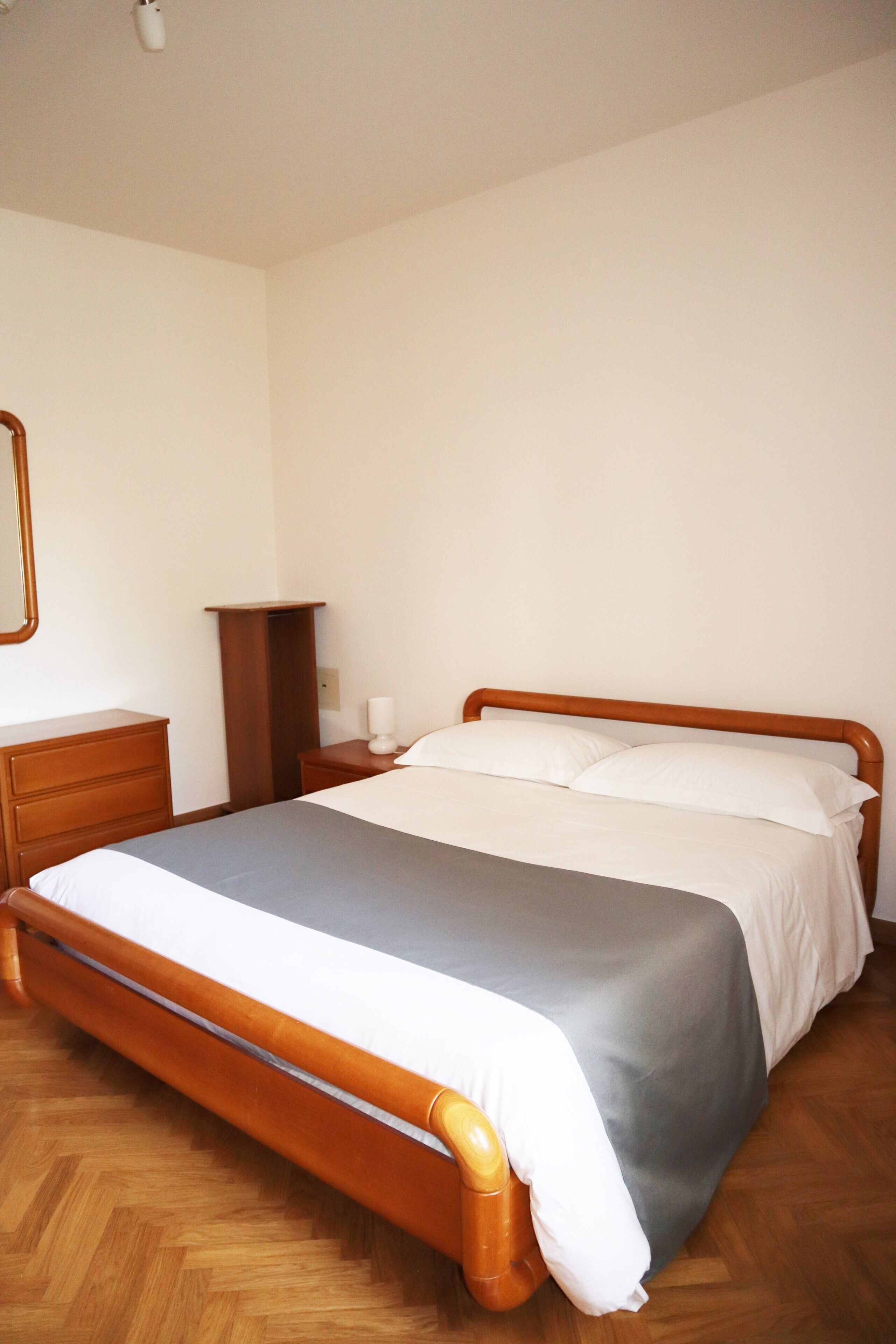 A neatly made bed with a wooden frame and grey bedding, a wooden nightstand with a lamp, and a wooden dresser with a mirror.