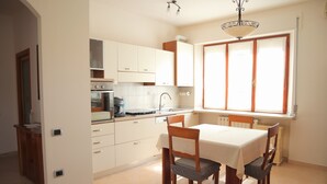 Une cuisine avec des meubles blancs, une table à manger avec des chaises et une fenêtre avec des stores.