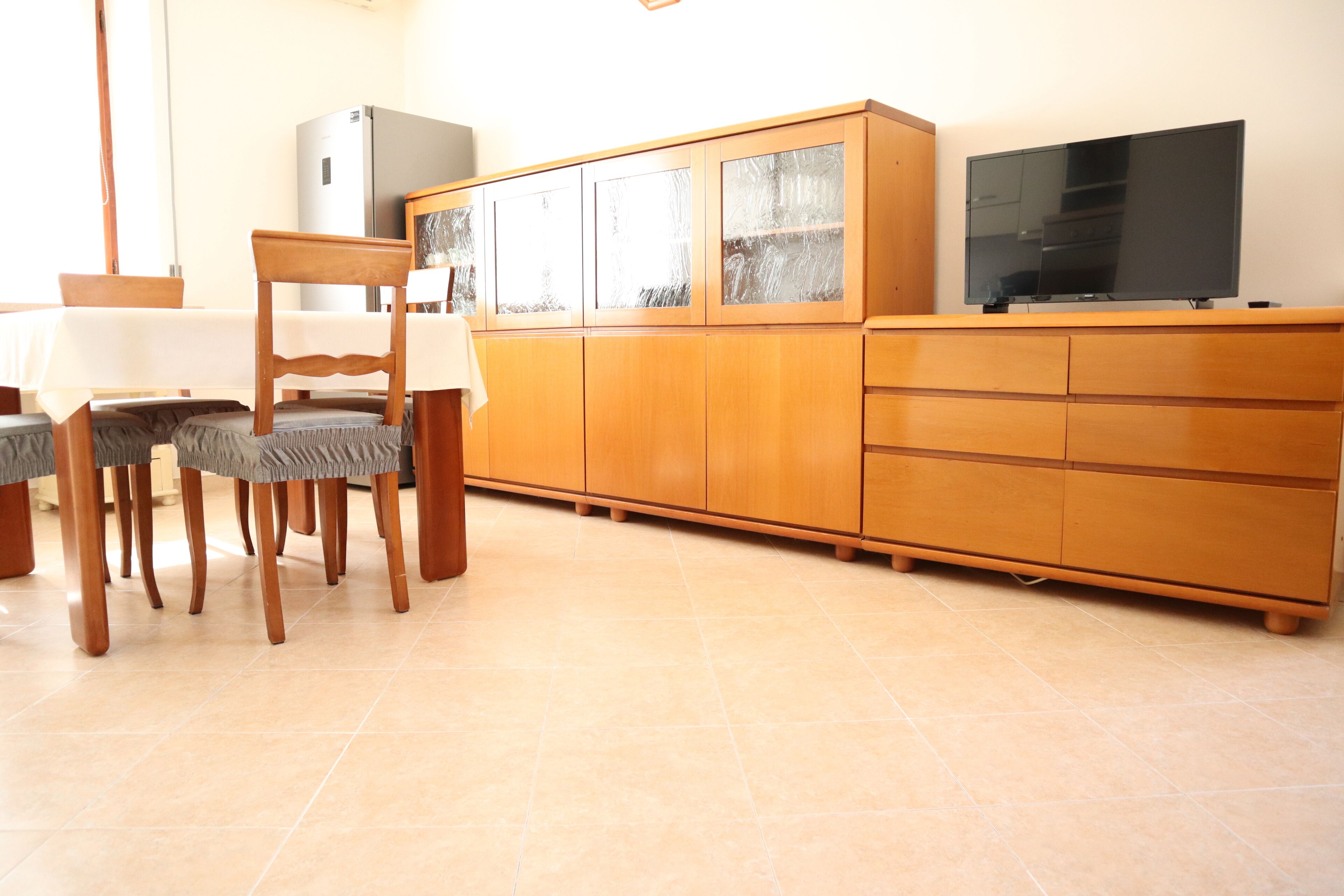 Una cocina con armarios de madera, un refrigerador, una mesa de comedor con sillas y un televisor sobre un mueble de madera.