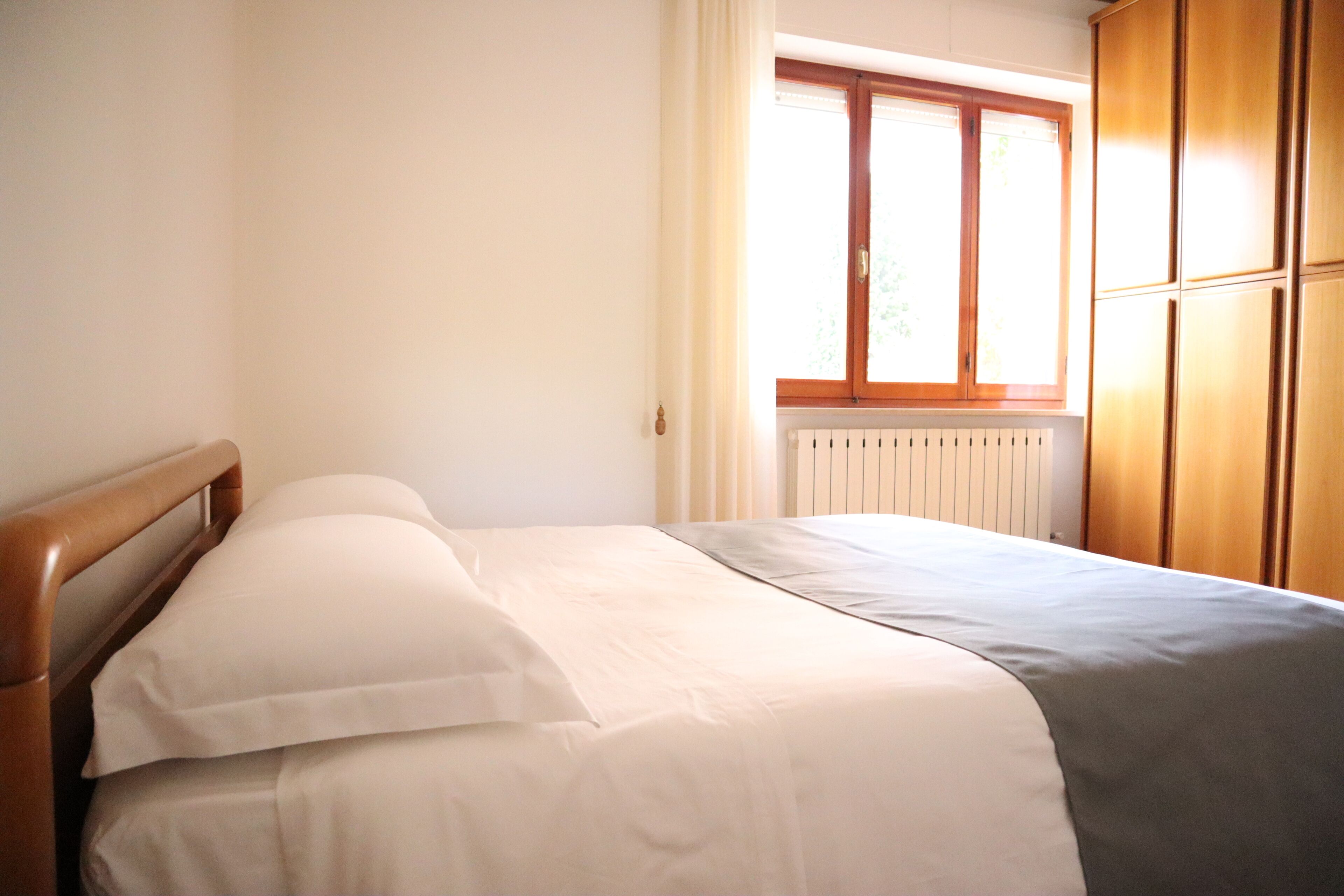 A bedroom with a wooden headboard, a bed with white and grey bedding, a window with curtains, and wooden wardrobe doors.