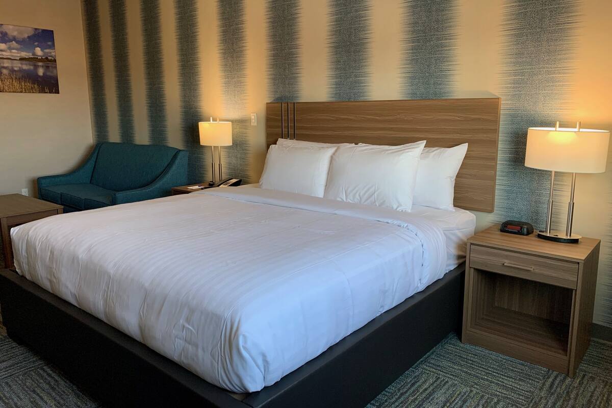 A hotel room with a large bed, two bedside tables with lamps, a wooden headboard, a teal armchair, and a painting on the wall.