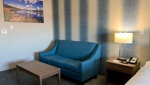 Quarto de hotel com um sofá azul, uma pequena mesa de madeira, uma mesa de cabeceira com abajur e telefone, e um quadro de um lago na parede.