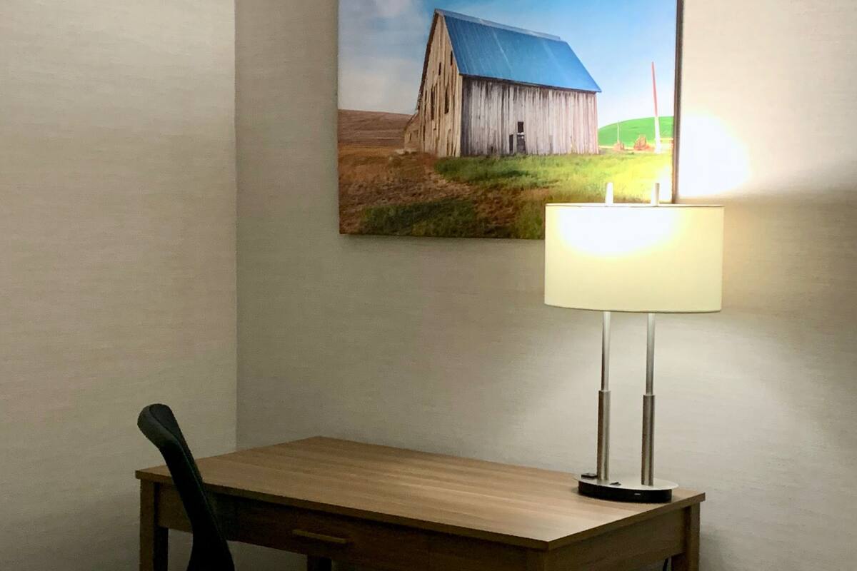 A hotel room with a wooden desk, a black office chair, a painting of a barn on the wall, and a table lamp.