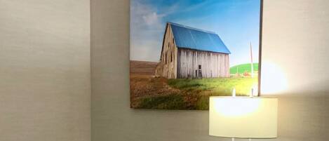 A hotel room with a wooden desk, a black office chair, a painting of a barn on the wall, and a table lamp.