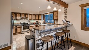 A modern kitchen with wooden cabinets, a large island with stools, stainless steel appliances, and a large window providing a view. - Mountain View Retreat - Epic Views, Pool Table & Garage (Rollinsville)