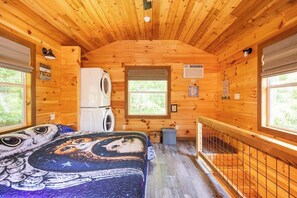 A wooden cabin interior with a bed, a washing machine, and a window. - Barn Owl Inn Treehouse - Private Hot Tub (Guild)