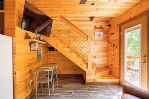 A wooden cabin interior with a TV mounted on the wall, a staircase, a seating area with a sofa, and a dining table with chairs.