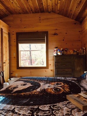 A wooden cabin room with a large window, a bed with a patterned blanket, and a cabinet with various items.
