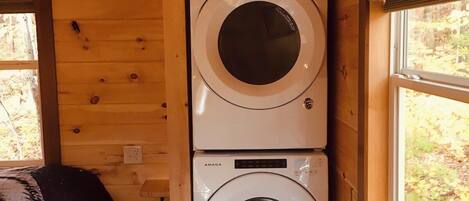 A laundry room with two stacked washing machines, a bed with a patterned blanket, and a window with a view of trees.