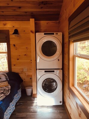 Un cuarto de lavado con dos lavadoras apiladas, una cama con una colcha estampada y un ventanal con vista a árboles.
