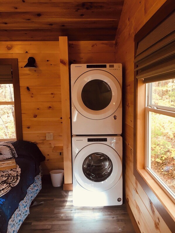 A laundry room with two stacked washing machines, a bed with a patterned blanket, and a window with a view of trees. - Barn Owl Inn Treehouse - Private Hot Tub (Guild)
