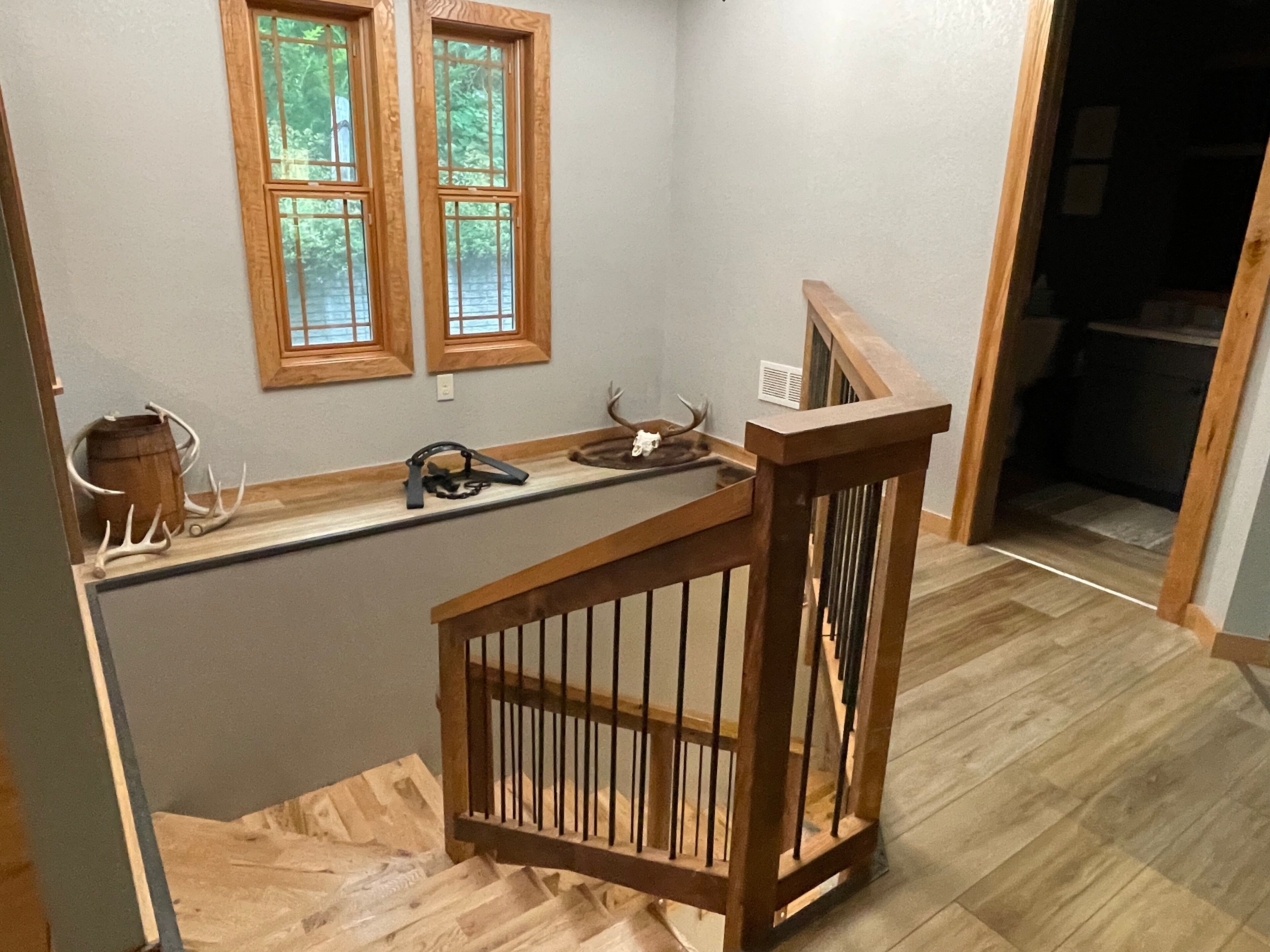 Una escalera de madera con barandilla, una ventana con vistas a la vegetación, y un estante con cuernos de ciervo y un barril.