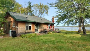 Eine Holzhütte mit grüner Dach, einem Grill und einer Bank am See.