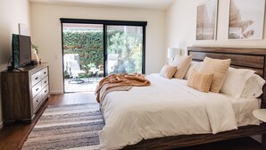 Ein Schlafzimmer mit einem großen Bett, einem Fernseher auf einer Holzkommode und einer Glasschiebetür, die auf eine Terrasse mit Gartenmöbeln führt.