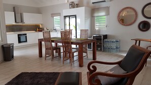 Un vaste espace de vie comprenant une table à manger et des chaises, un canapé et une table basse. Il y a également un coin cuisine avec des meubles blancs et un four encastré. La pièce est dotée d’un haut plafond avec des poutres apparentes et d’une grande fenêtre qui laisse entrer la lumière naturelle.
