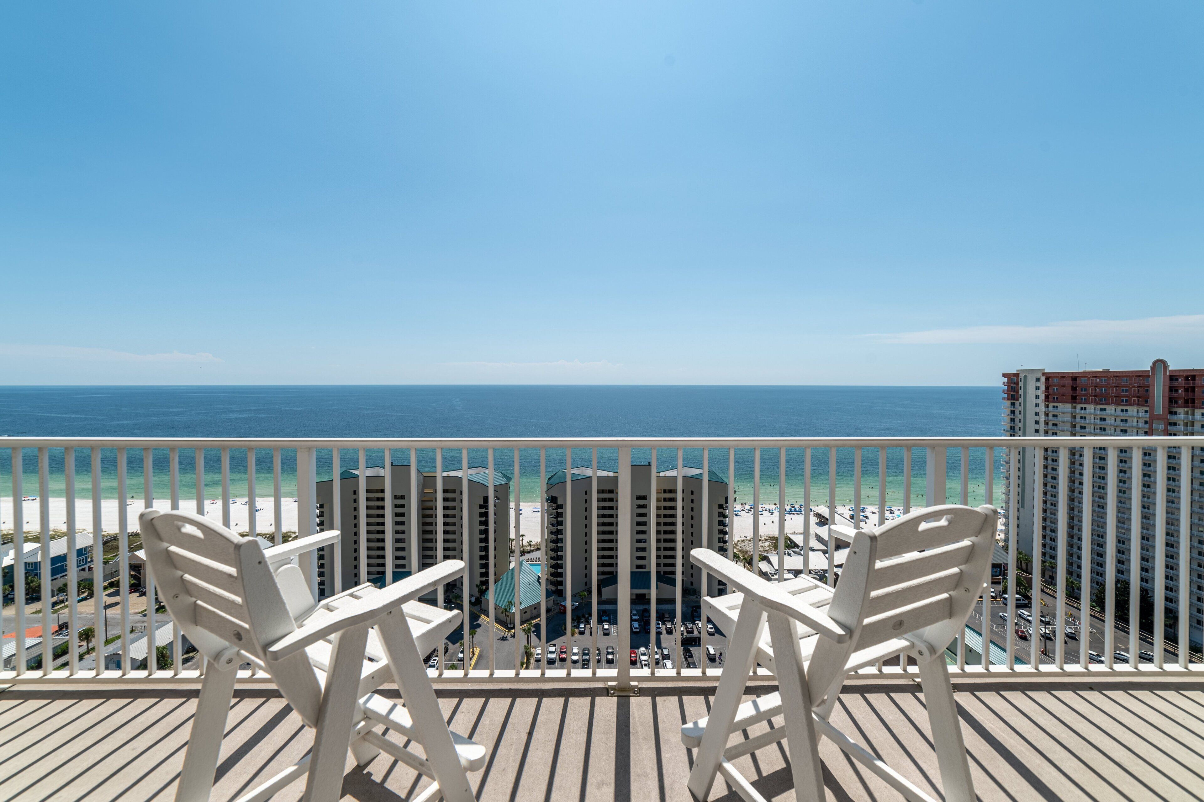A balcony with two white chairs overlooking a beach and a pool area, with buildings in the background.