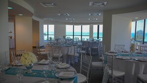 Ein Festsaal mit runden Tischen, gedeckt für eine festliche Veranstaltung, große Fenster mit Blick auf das Meer und ein modernes Decken­design.