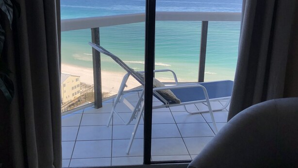 Ein Zimmer mit großem Fenster mit Blick auf Strand und Meer. Im Vordergrund stehen ein Sessel und ein kleiner Tisch.