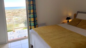 Un dormitorio con cama, mesita de noche con lámpara, vista al mar a través de una ventana y piso con estampado.
