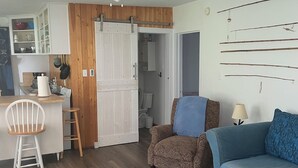 Ett mysigt vardagsrum med trägolv, en blå soffa, ett träbord och en vit stol. Det finns också en köksdel med ett vitt skåp och en trädörr.