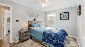 A bedroom with a bed, nightstand, and ceiling fan. There is a window with blinds and a door leading to another room. - PET FRIENDLY - MINUTES TO PARRIS ISLAND - BEACHES (Beaufort)