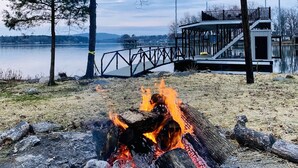 A lakeside view with a fire pit, a wooden deck, and a distant bridge.