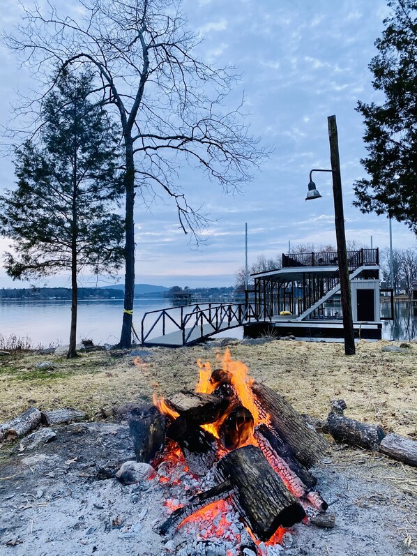 A lakeside view with a fire pit, a wooden deck, and a distant bridge.