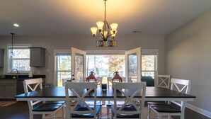 A dining area with a dark wooden table and white chairs, a chandelier, and a view of trees through large windows.