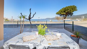 Area esterna con zona pranzo, tavolo apparecchiato per due, sedie, una pianta in vaso e vista sul mare e sul porto turistico.