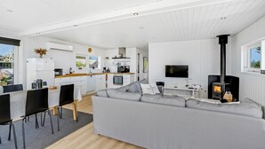 A modern, white-themed living room with a grey sofa, a dining area with a table and chairs, a kitchen with white cabinets and a stove, and a TV mounted on the wall.
