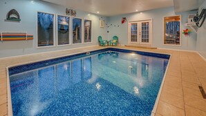 Ein Innenpool mit klarem, blauem Wasser, umgeben von beigen Fliesen. In der Nähe des Pools befinden sich zwei grüne Stühle und ein kleiner Tisch, und ein Fenster mit Blick auf Bäume.