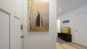 Un couloir avec une porte blanche, un tableau représentant des bouleaux, un meuble noir pour appareils électroniques et un téléviseur affichant une image d’un arbre jaune.