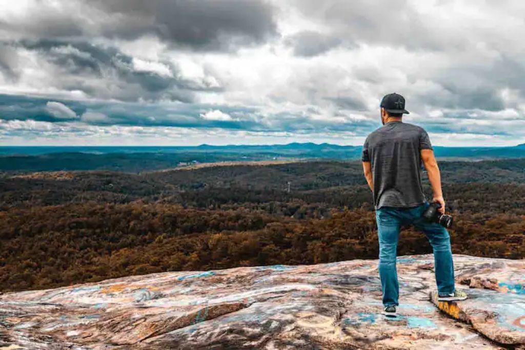 A man standing on a rock with a camera, overlooking a forested landscape.