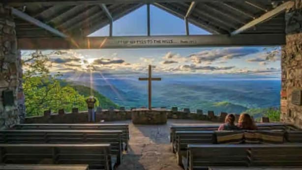 Innenansicht einer Kirche mit Kirchenbänken, einem Kruzifix und Blick auf eine Landschaft durch ein großes Fenster.