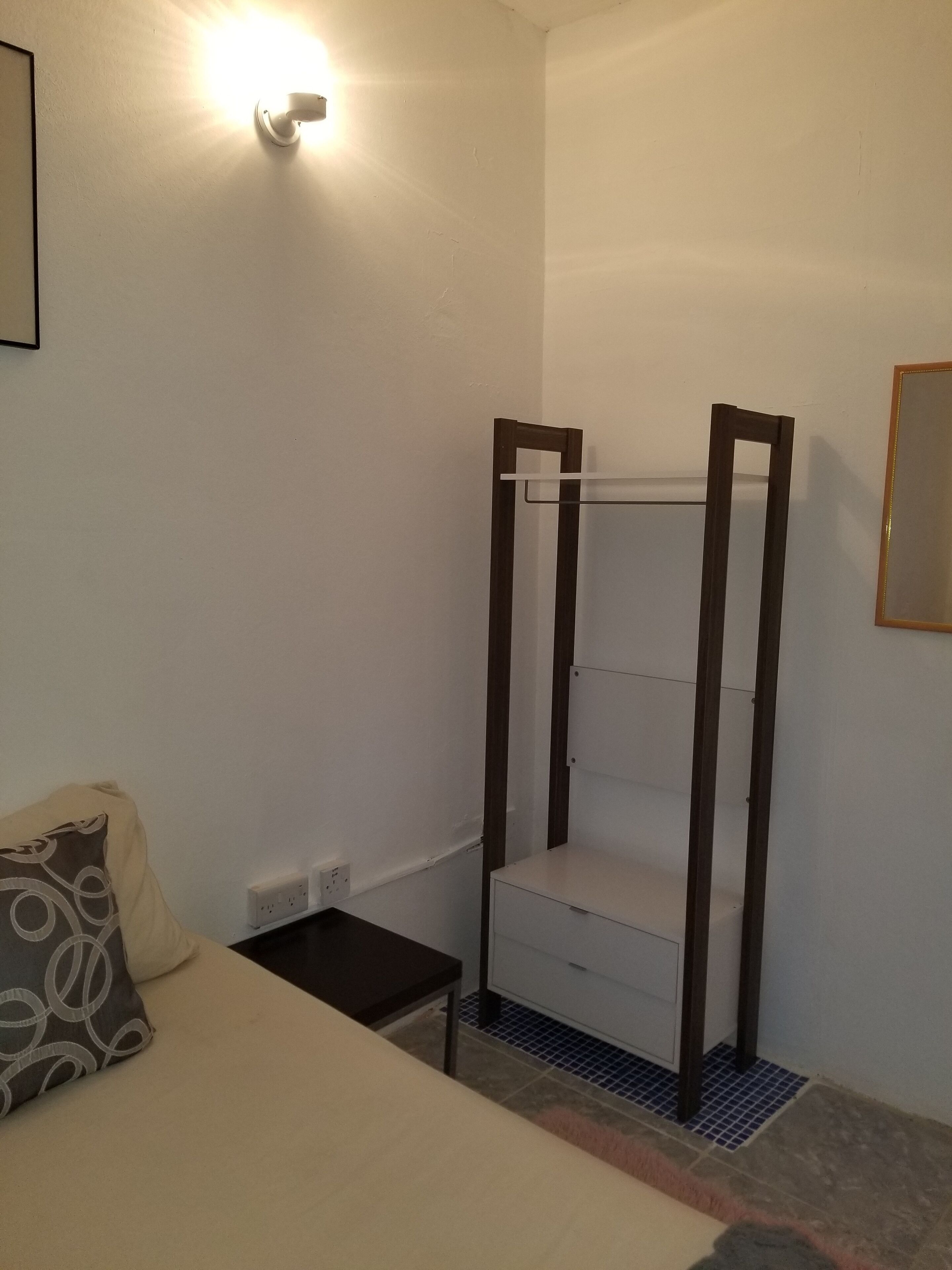 Una habitación pequeña y sencilla con una cama, una mesita de noche, un estante en la pared y un espejo.