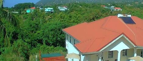 A large, two-story building with a red roof and white walls, surrounded by lush greenery and mountains in the background.