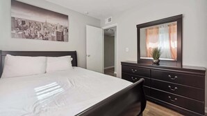 A bedroom with a bed, a dresser, a mirror, and a cityscape painting on the wall. - Fairlane #6 | Classy Cool & Chic (Lansing)
