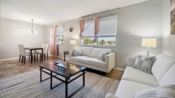 A living room with a sofa, coffee table, dining table, chairs, and a view through the window. - Fairlane #6 | Classy Cool & Chic (Lansing)