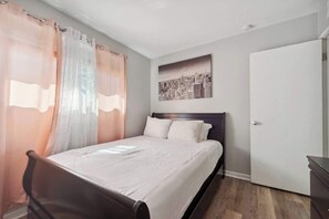 A bedroom with a bed, curtains, a cityscape painting, a wardrobe, and a door. - Fairlane #6 | Classy Cool & Chic (Lansing)