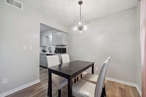 A modern dining area with a dark wooden table and white chairs, a kitchen in the background, and a unique ceiling light fixture. - Fairlane #6 | Classy Cool & Chic (Lansing)