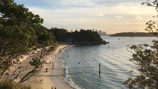 A beach with people walking along the shore, a body of water, and a city skyline in the background.
