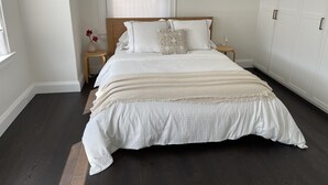 A bedroom with a large bed, wooden headboard, white bedding, a bedside table with a lamp, and a window with blinds.