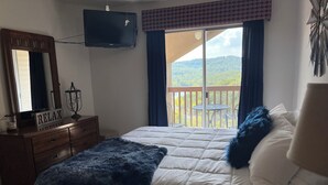 A bedroom with a bed, a dresser, a television, and a balcony with a view of the outdoors.