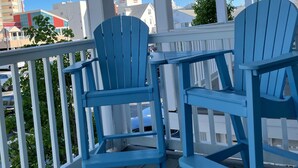 Une véranda couverte avec deux fauteuils Adirondack bleus, des balustrades blanches et une vue sur les bâtiments en arrière-plan.